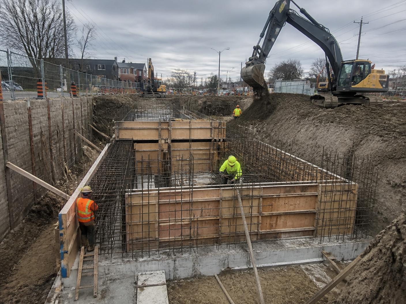 Cast-in-place concrete civil structure under construction in a shored excavation with excavator and crew